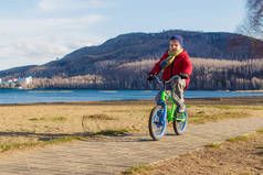 春天, 在山海的背景下, 6岁的男孩穿着夹克、围巾和帽子, 骑着两轮儿童自行车在人行道上