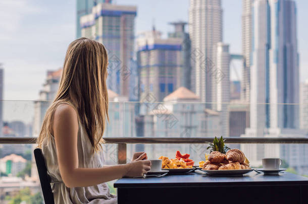 一个年轻的女人正在阳台上吃<strong>早饭</strong>。早餐桌与咖啡果子和面包 croisant 在阳台反对大都市的背景