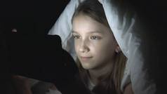 儿童玩平板电脑在黑暗的夜晚, 女孩浏览互联网在床上, 而不是睡觉
