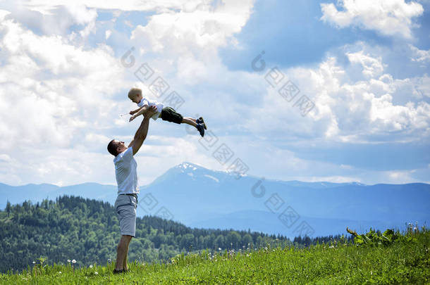 快乐的父亲在绿色森林、山脉和天空的背景下, 把一个小儿子扔了出来。亲子友谊