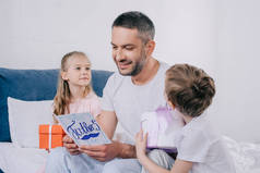 可爱的女儿和儿子坐在礼品盒附近微笑爸爸拿着父亲节贺卡