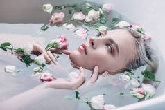 美丽的女人躺在清澈的水里，花在白色的浴缸里，看着远方