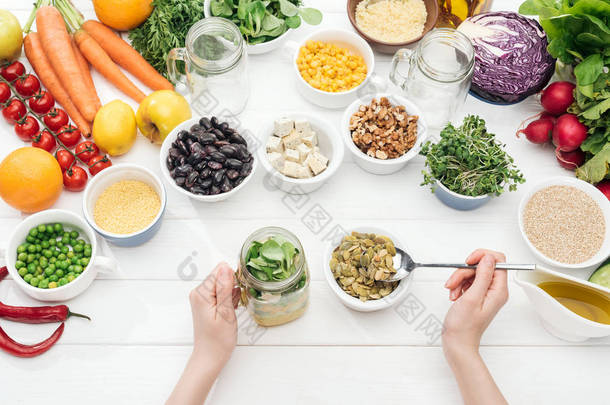 裁剪视图的妇女添加南瓜种子在罐子与沙拉在木制白色桌子上