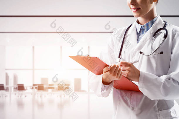 亲密的自信的女医生穿着白色的医疗制服, 一边站在明亮的医院<strong>大楼内</strong>, 一边把笔记写进笔记本。医疗行业理念