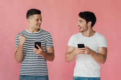 两个微笑的男人使用智能手机,看着对方孤立的粉红色
