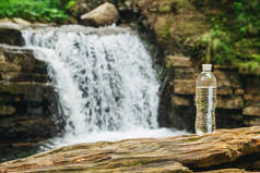 透明的塑料瓶与水倒立在一个木制树干上，面对着河流和瀑布的背景。纯天然饮用水的概念