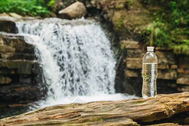 透明的塑料瓶与水倒立在一个木制树干上，面对着河流和瀑布的背景。<strong>纯天然</strong>饮用水的概念
