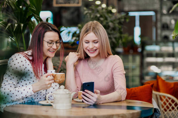 两个年轻的漂亮女孩坐在一张绿色的咖啡馆里。喝羊角面包喝茶，聊天，大笑，在智能手机上拍照.