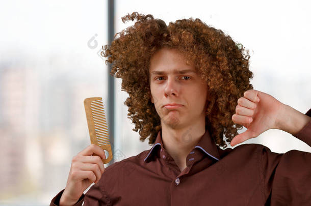 画像一个卷发的年轻人，一个木梳在宏伟的头发不满的结果对窗口。男性头发护理概念