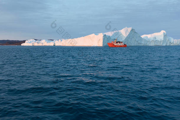 冰山之间的小船。在格陵兰岛伊卢利萨特的午夜太阳期间，帆船在迪斯科湾冰川漂浮的冰山之间巡航。全球变暖冰和冰山现象的研究