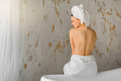 美丽的年轻女子在白色毛巾在她的头上坐在她的背部在浴室