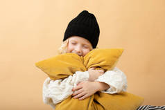 一个身穿白色针织连衣裙、头戴黑色帽子的小女孩坐在黄色背景的格子布上。 女孩抱着橙色枕头