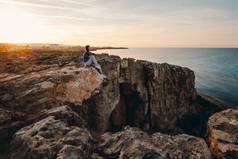 在夏天的群山中，女旅行家坐在悬崖顶上的海边，欣赏着大海和大自然的美景。 希腊角，塞浦路斯，地中海。 日出