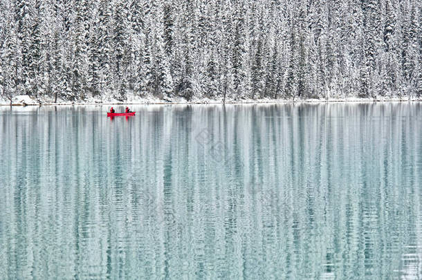 两个人乘红船在平静的蓝色湖上划船，周围是覆盖着积雪的冬季森林。 美丽的冬季森林和路易斯湖中的红色独木舟的映照。 加拿大落基山脉 班夫国家公园 艾伯塔省 加拿大.