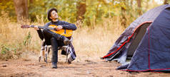 带着黑帽微笑的年轻人坐在旅游帐篷附近，在森林里弹吉他的横幅图案