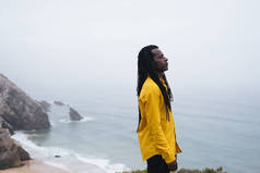 身披长发的非洲男性站在海岸上，白浪和悬崖峭壁笼罩着背景