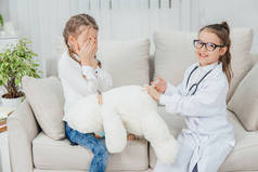 穿着医生制服的可爱小女孩正在给白泰迪熊打针。 另一个女孩正坐在那里，满脸忧心忡忡，双手捂住脸.