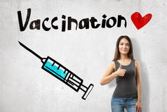 一个年轻的微笑的女孩，带着注射器的形象和可爱的小红心疫苗的称号，在墙边竖起大拇指.
