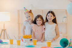 快乐的三个女孩的室内镜头拥抱和玩乐，开心的笑着，站在节庆桌旁，端着蛋糕、杯子，有着快乐的童年，在一起聚会。童年和节日概念