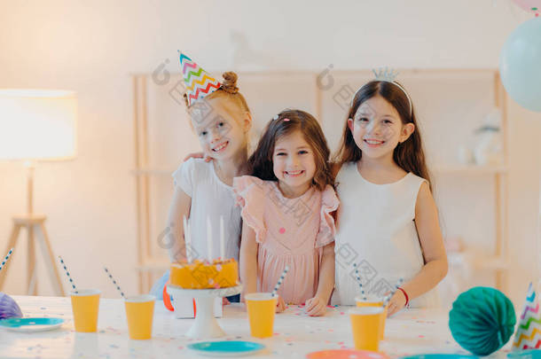 快乐的三个女孩的室内镜头拥抱和玩乐，开心的笑着，站在节庆桌旁，端着蛋糕、杯子，有着快乐的<strong>童年</strong>，在一起聚会。<strong>童年</strong>和节日概念
