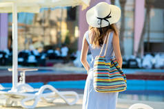 夏日晴天，身穿浅色夏装，头戴黄色草帽，头戴时尚肩袋的年轻女子站在酒店门外.