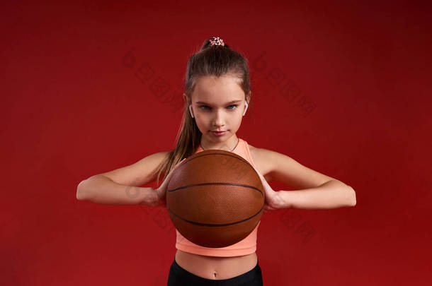 保持动力。一个可爱的孩子，一个从事体育运动的女孩，她一边打篮球一边看着镜头。被红色背景隔离。健康，训练，积极的生活方式概念。<strong>横向</strong>射击