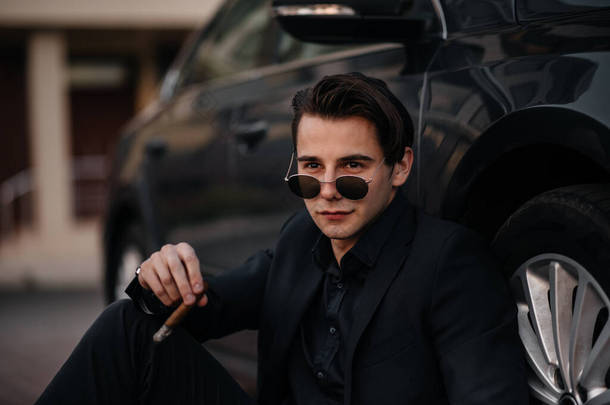 一个时髦的商人在一辆豪华汽车旁边抽烟雪茄.时尚与商业