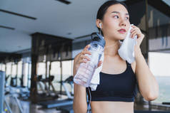 亚洲健康女性运动后在健身房放松，手握纯净水和白毛巾