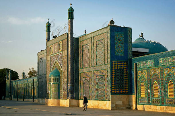 阿富汗巴尔赫省马扎里沙里夫。一名男子在<strong>阳光下</strong>走过马扎里沙里夫的蓝色清真寺。多彩的马赛克和瓷砖装饰着清真寺的墙壁。阿富汗北部.