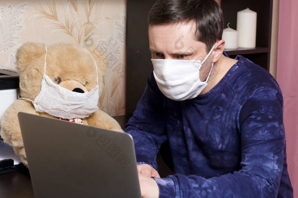 一个戴防护纱布面具的男人在笔记本电脑后面，在自我隔离的条件下工作。附近有一个柔软的玩具。熊戴上防护罩.