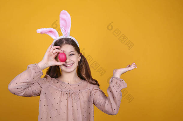 画室里的年轻姑娘头戴传统的<strong>兔子</strong>耳朵帽带为复活节，用鸡蛋盖住她的<strong>眼睛</strong>，面带微笑。黄底上有辫子的布鲁内特雌性。关闭，复制空间.