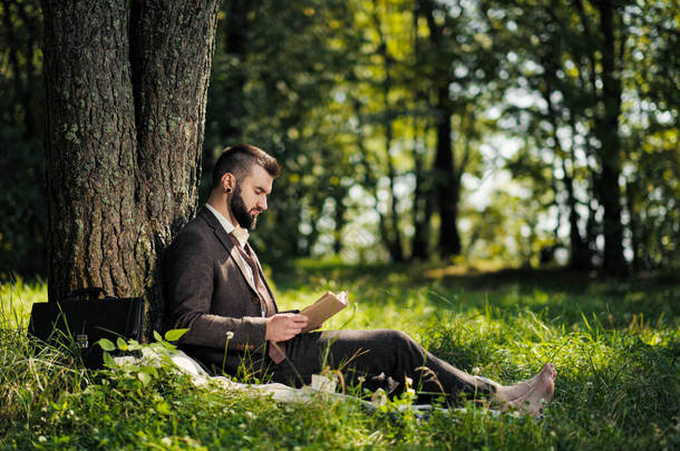 年轻迷人的胡子商人坐在树下的绿草上，在公园里休息。读书，喝咖啡。放松，厌倦了工作，午休.