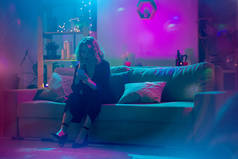 穿着黑色雅致西服的金发女青年坐在柔软的沙发上，在被粉色和蓝色灯光照亮的客厅里用智能手机打滚