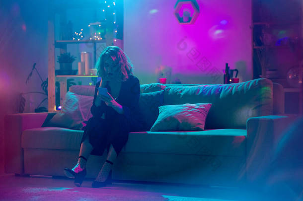 穿着黑色雅致西服的金发女青年坐在柔软的沙发上，在被粉色和蓝色灯光照亮的客厅里用智能手机打滚