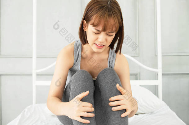 年轻美丽的亚洲女人，穿着灰色的衣服，早上醒来，躺在一张白色的床上，身体疼痛，双手抱在肚子上，腹部疼痛，胃痛，疼痛，疼痛