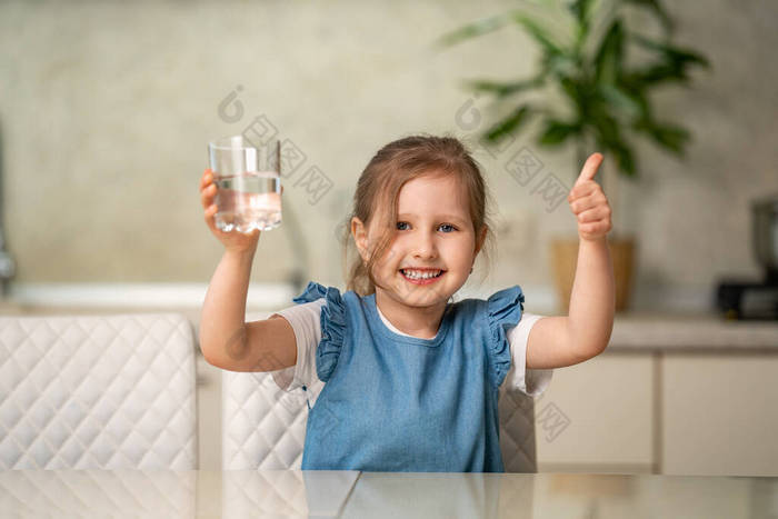 可爱的小女孩在家里厨房里喝水。小女孩高兴地笑着，手里拿着一杯清澈的水，表示赞同。水平衡。防止脱水