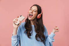 在粉色背景下使用耳机和手机唱歌的年轻女子的照片