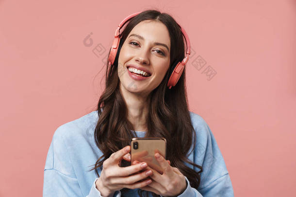 在粉色背景下使用耳机和手机时高兴地大笑的年轻女性的照片