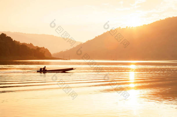 亚洲的男性渔民在黄昏的时候驾驶木船在湖上钓鱼，夕阳在山后的背景下，湖面上闪烁着明亮的光芒，在泰国北部的乡村风光.