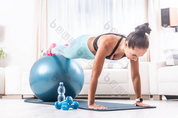 年轻的女运动员在客厅里保持着一块木板的姿势，她的腿在健身球的顶部，还有一瓶水、两个哑铃和一个垫子.