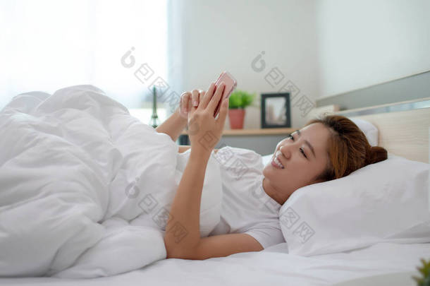 亚洲女人正在卧室的床上玩智能手机。她睡觉前感到很放松.