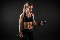 健身、运动和健康的生活方式理念.漂亮的女运动员在黑色背景上展示她受过训练的身体。健美女子模特儿用哑铃训练二头肌.