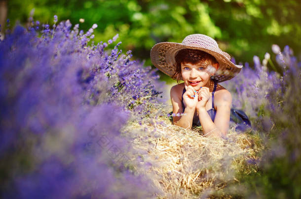 可爱的微笑的小女孩，身穿蓝色的衣服，头戴白色的帽子，躺在薰衣草地里，夏日阳光灿烂，周围开着紫罗兰的花朵。普罗旺斯的夏天旅行。薰衣草中的儿童