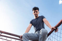 西班牙裔年轻人坐在屋顶上观看日落- -男人坐在蓝天的旧金属网架上- -都市风格