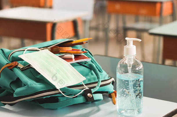 COVID-19预防、重返校园和新的正常观念。在课堂课桌上放有学习用品的背包里放有清洁剂凝胶和外科口罩的前景.