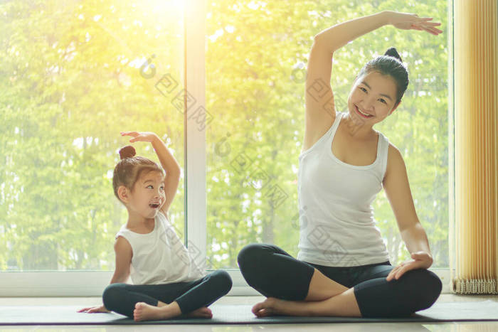 亚洲妈妈在家里练习瑜伽，身边坐着一个可爱的女儿，一边在家里锻炼，一边笑着模仿妈妈的姿势.