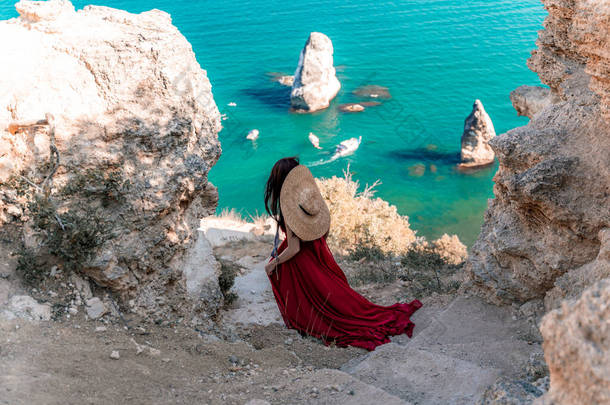 从后面看去，一个女人穿着红色的飞行服，在风中飘扬。戴着草帽，带着岩石和小船在<strong>大海</strong>的<strong>背景</strong>下走下楼梯。浪漫的概念