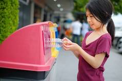 一张特写照片，是一个可爱的年轻亚洲女孩把一个空的塑料水瓶扔进了一个回收箱，回收箱将废物分类为3组，回收瓶、回收罐、纸张和垃圾.