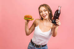 年轻美丽饥饿的女人吃汉堡包的画像。具有粉色背景的快餐学生的孤立肖像。饮食概念.