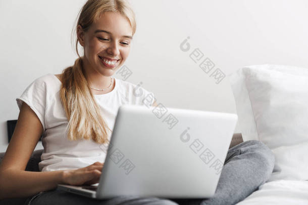 照片中快乐的年轻女性坐在明亮的房间里，一边笑着一边用笔记本电脑工作